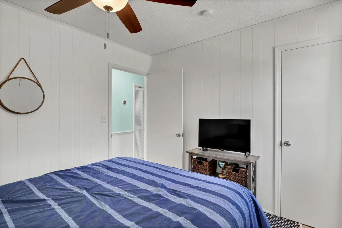 A calm and comfortable bedroom with a striped blue bedspread, wall-mounted TV, decorative mirror, and open doorways. Sunlight softly brightens the room, creating a peaceful atmosphere ideal for relaxation.