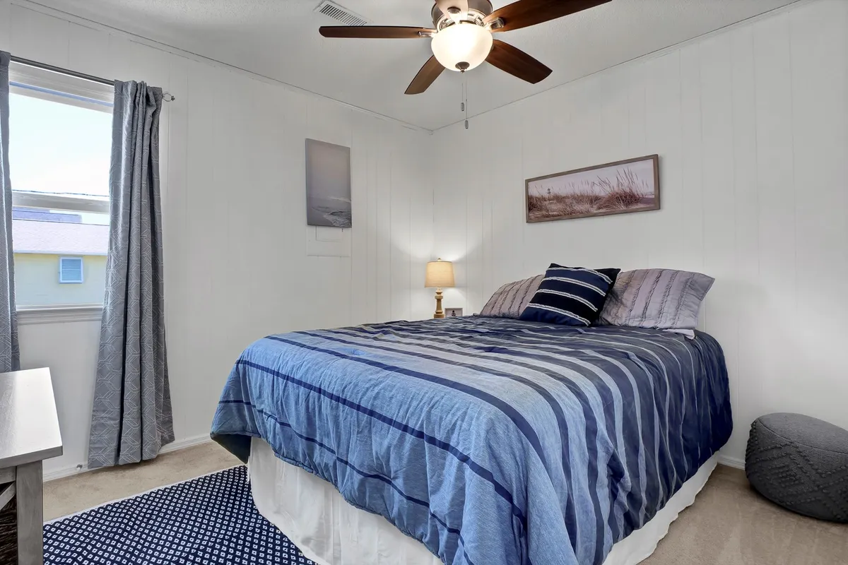 A relaxed bedroom featuring a bed with blue striped bedding, a table lamp, a window with curtains, and framed wall art. The space feels intimate and peaceful, inviting restful sleep after a day of coastal adventures.