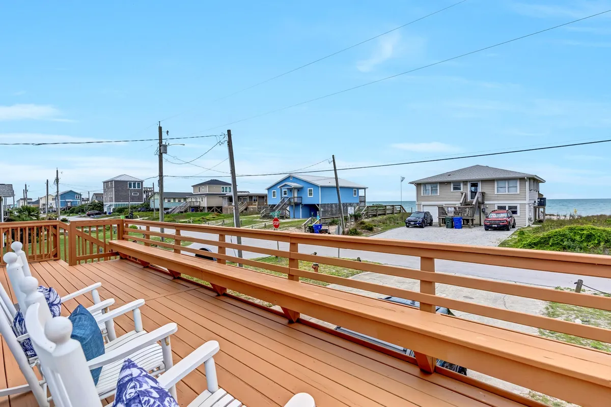 The image captures the inviting exterior deck of a single family house, showcasing classic white deck railings and a row of cozy rocking chairs facing the ocean. The view is dominated by the sparkling water and expansive sky at sunset, with gentle ocean breezes creating a relaxed, coastal ambiance. Neighboring beach houses are visible, highlighting a sense of community and beachside living.