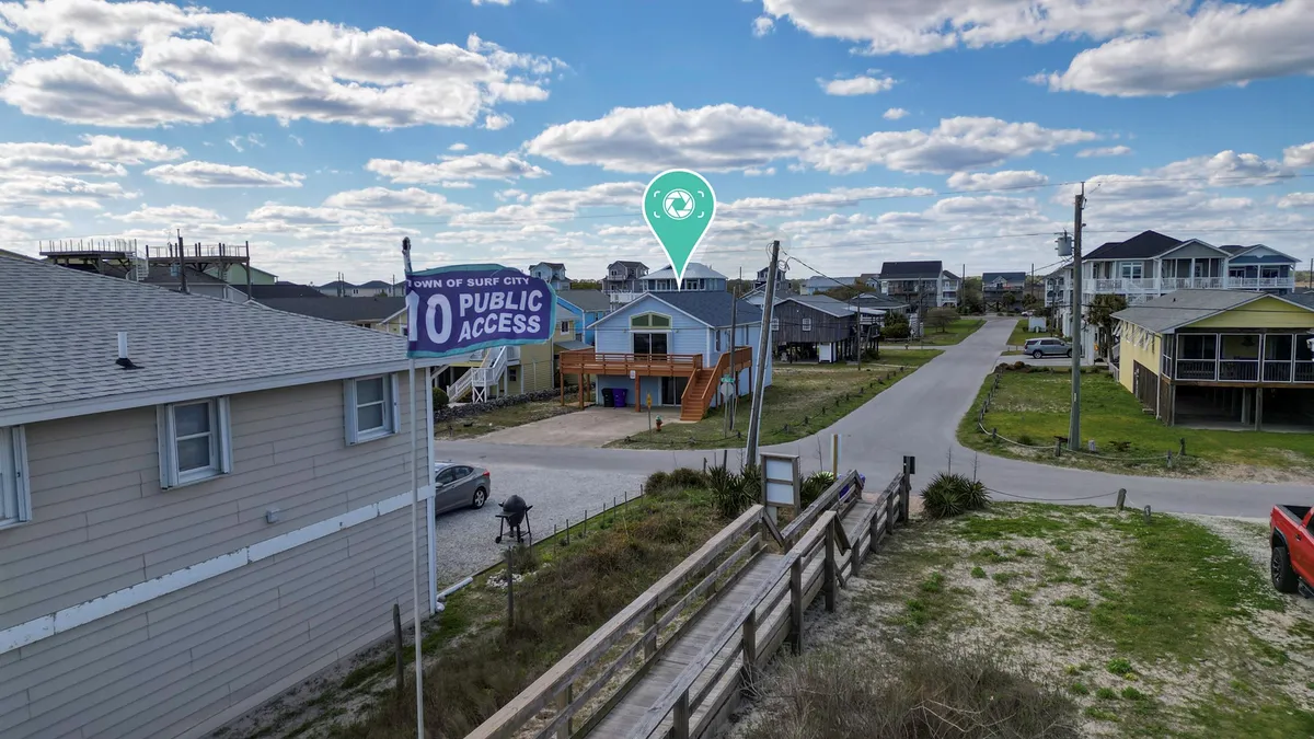 An exterior shot highlighting a public access sign, a blue house, and wooden steps leading down, set against a relaxed beach-town backdrop. The scene suggests both community and ease of access to outdoor activities.