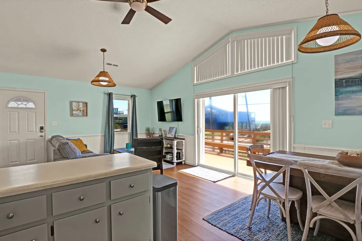 An open living and dining space with a kitchen island, a dining table, a sliding glass door leading to the deck, and a comfortable living room seating area. Natural light fills the room, and the arrangement facilitates easy movement and togetherness.