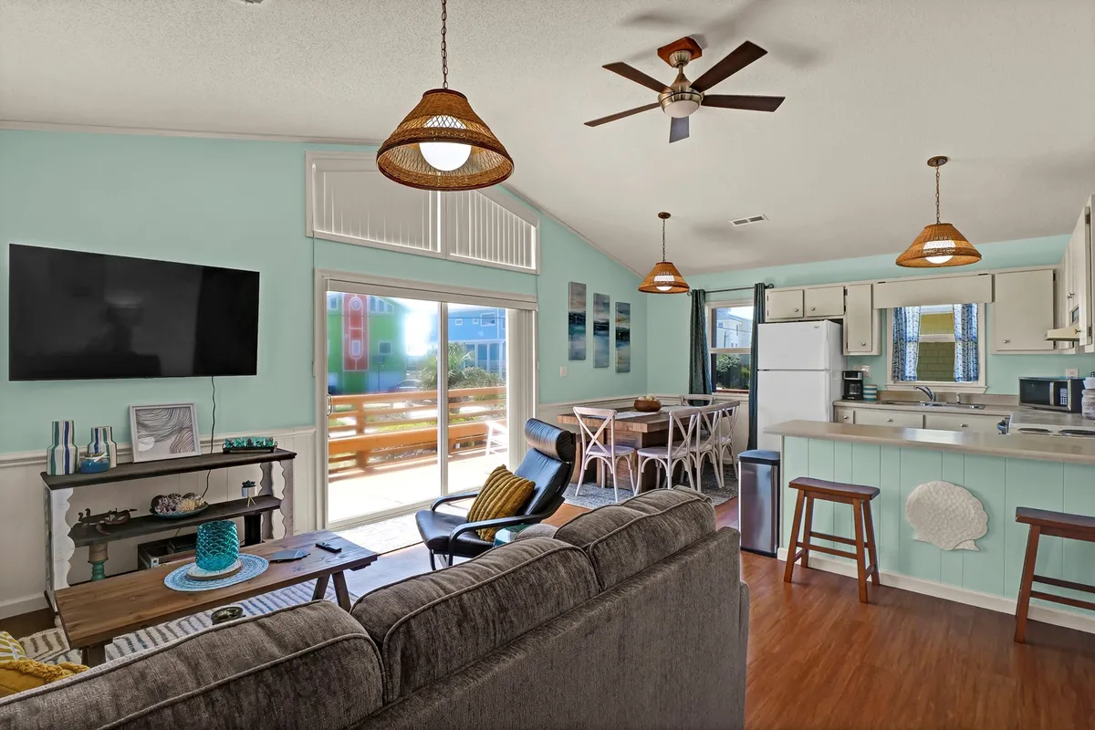 A casual and inviting interior scene featuring a comfortable sofa, a dining table, a kitchen area, and large windows that let in plenty of natural light. The space feels open, with a focus on shared meals and relaxation after beach days.